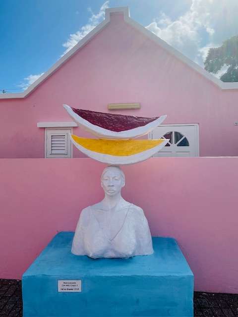 White statue of woman with two large slices of fruit on her head on a blue pedestal in front of pink cottage and fence in Curacao