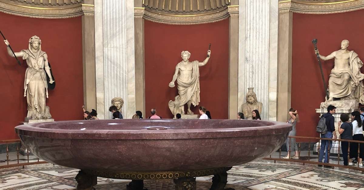 A trio of statues in front of a red background with a burgundy colored bath in the foreground at The Vatican Museum in Rome