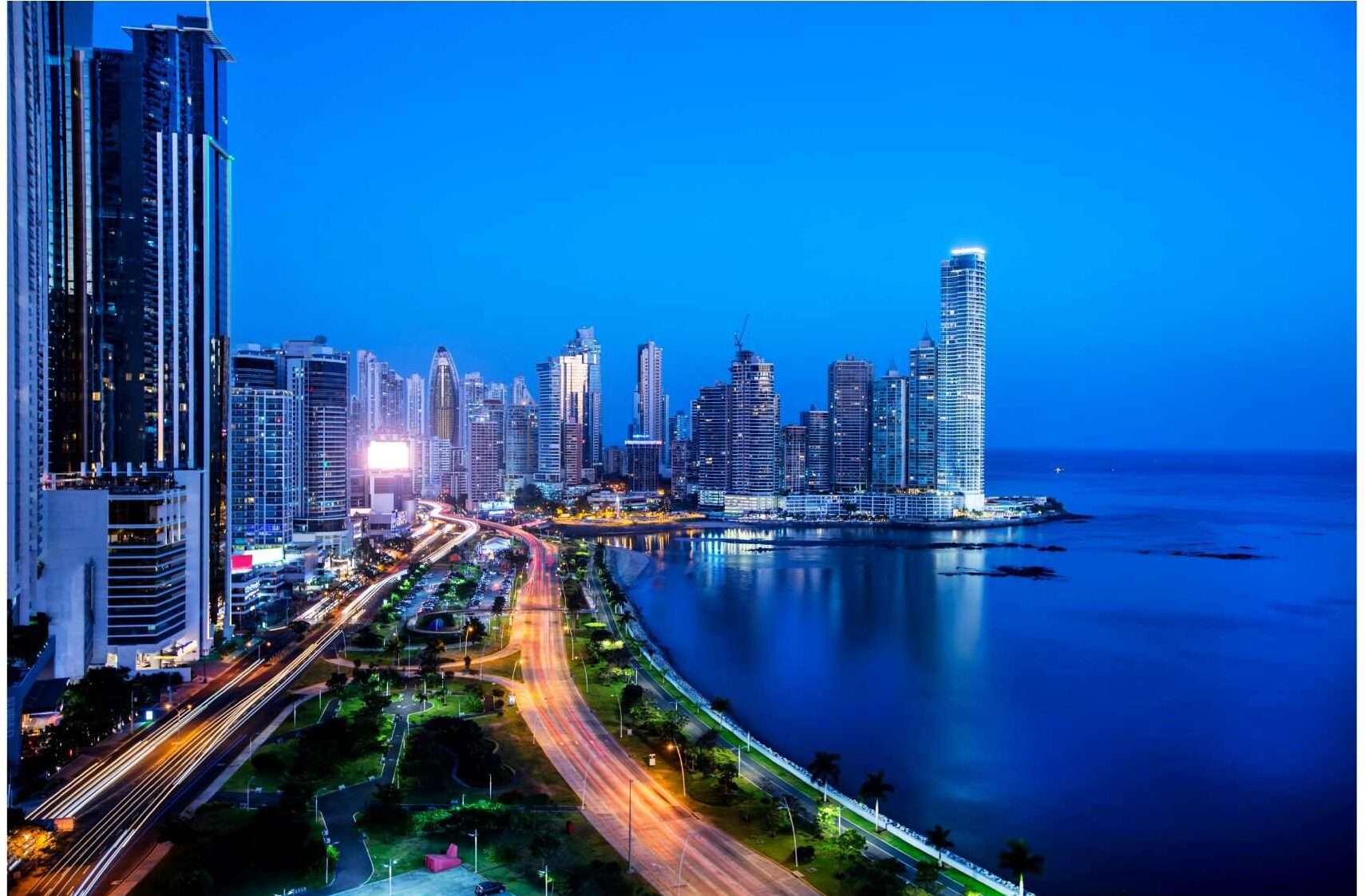 Many skyscrapers with evening lights on sit next to the well-lit waterfront highway at dusk in Panama City
