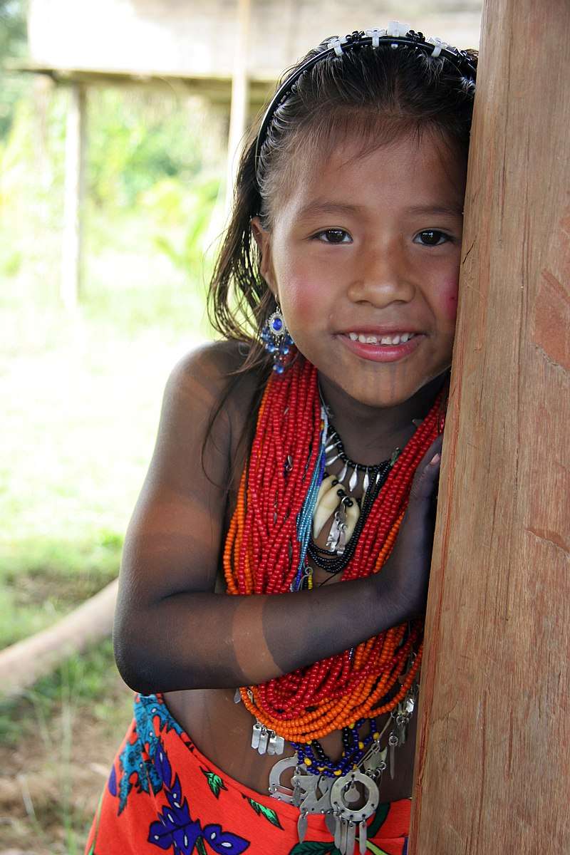 Shy but smiling young girl wearing colorful orange and blue jewelry as she leans against the tree