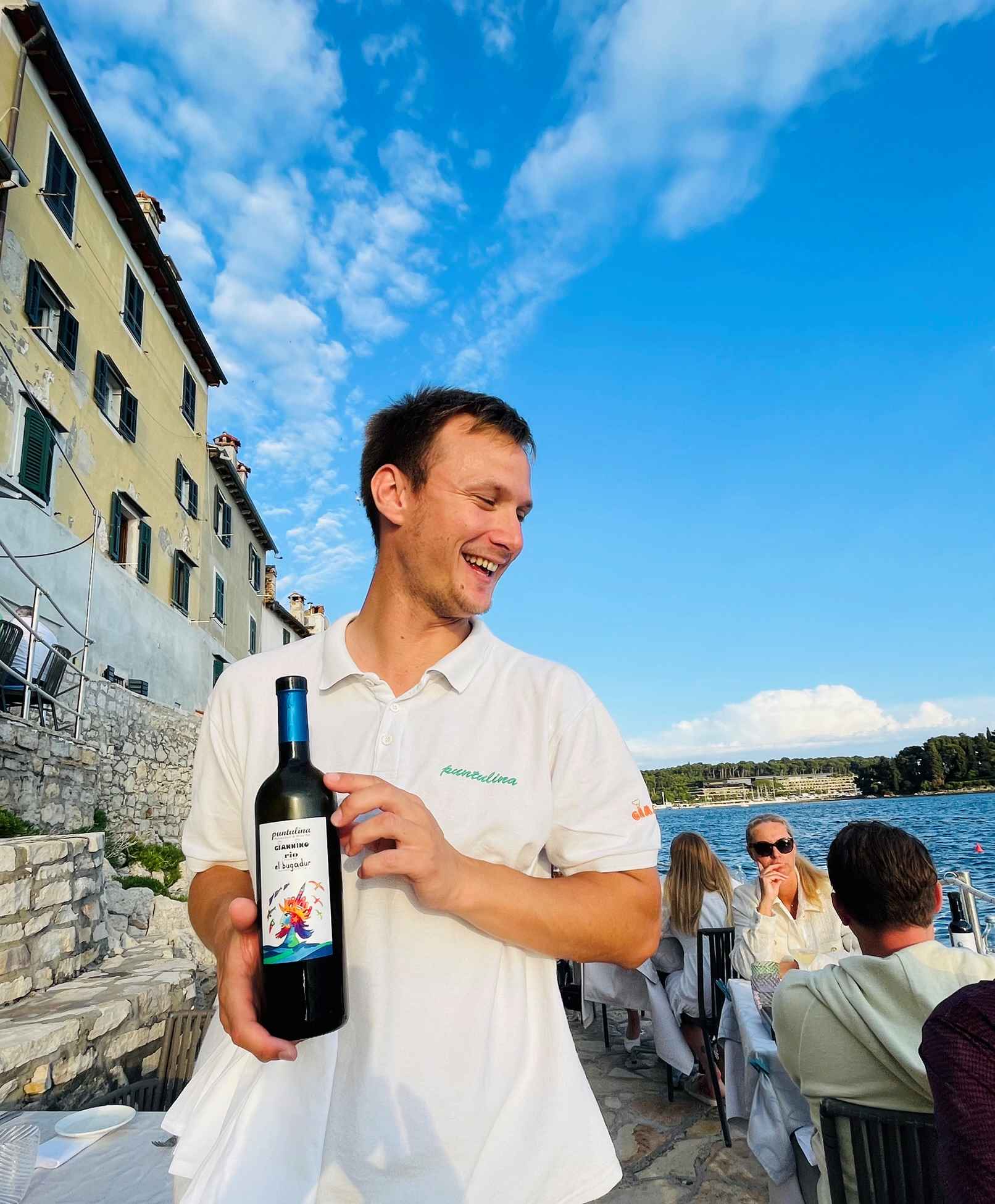 Smiling waiter in white uniform holding a bottle of wine on the rocks in Rovinj, Croatia