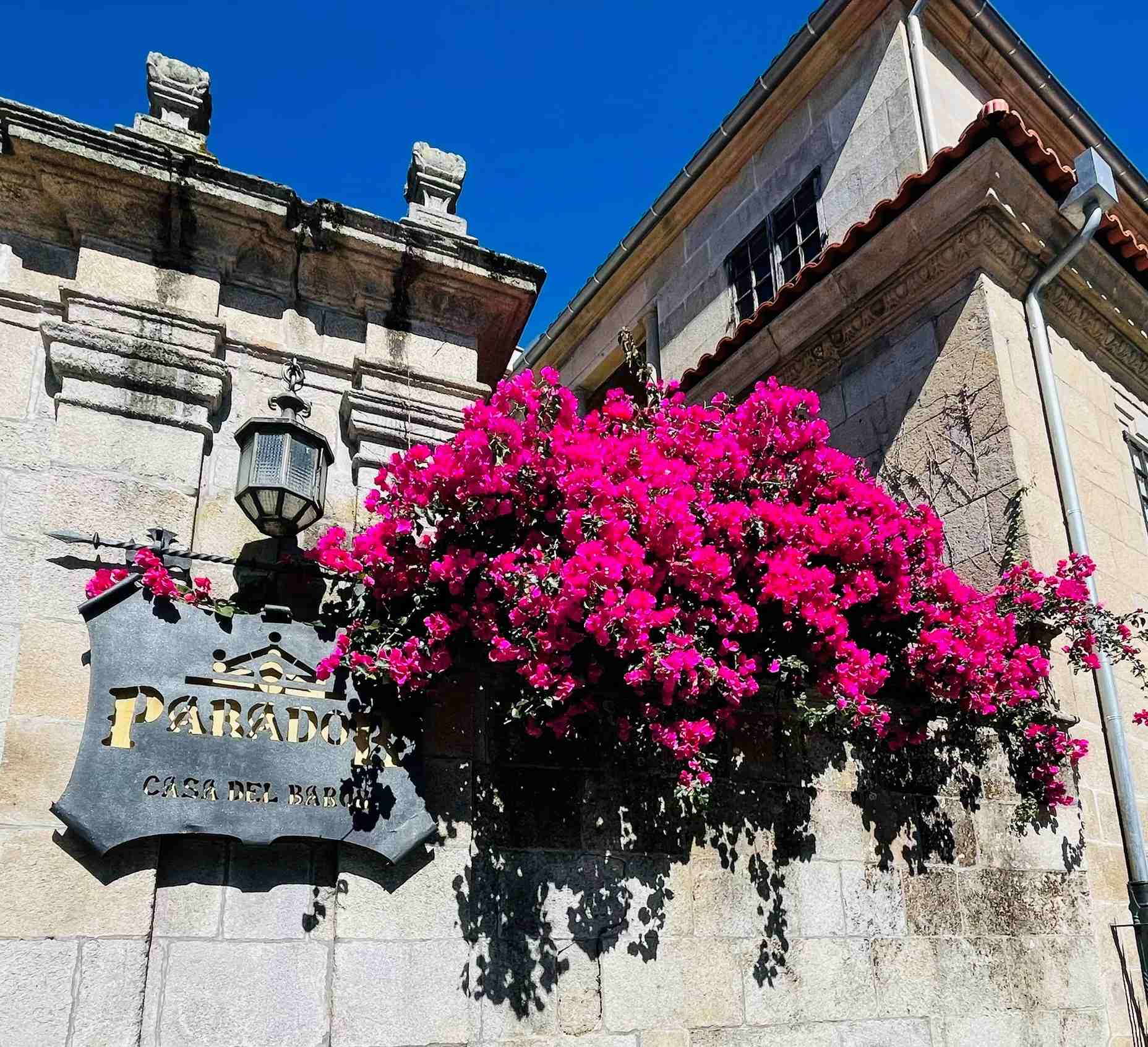 Cascading bright pink flowers against stone building and a sign saying Paradore Casa Del Baron in Ponte Vedra, Spain