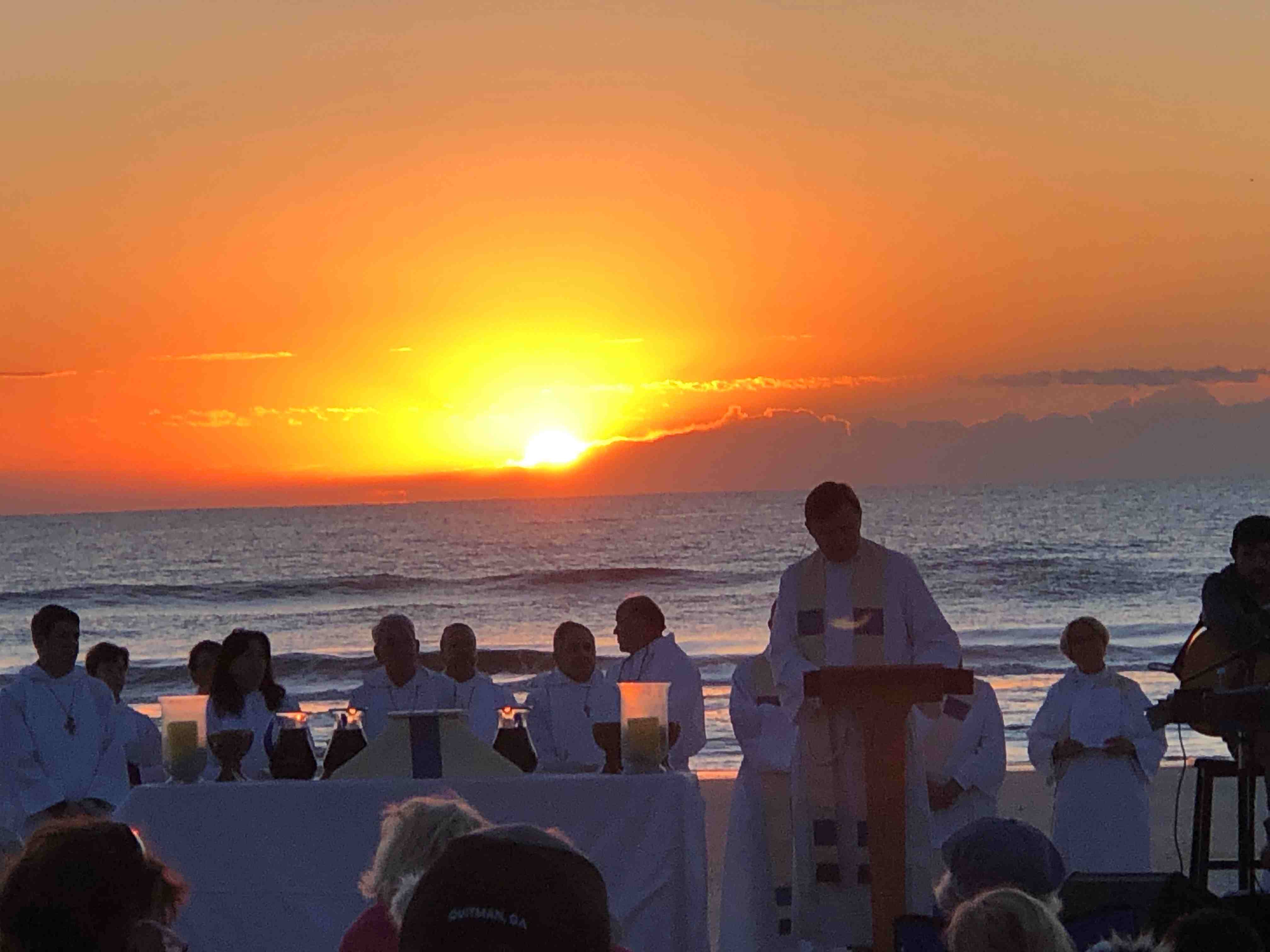 Orange sunrise over Atlantic Ocean with many ministers dressed in white as they prepare to give communion to parisioners