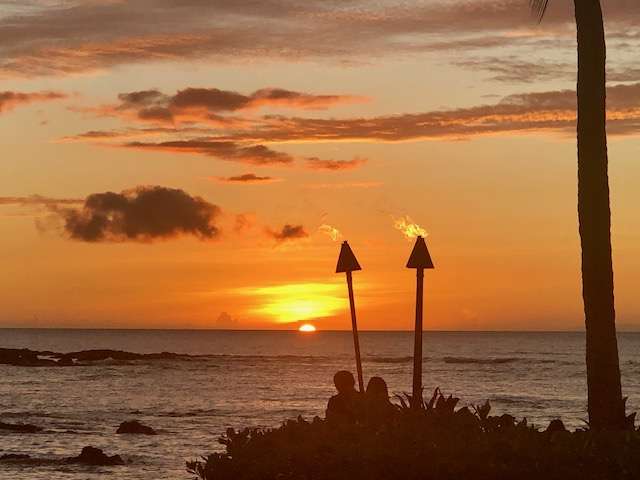 A couple watches the dreamy gray clouds and orange sky at sunset with tiki torches in foreground