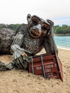 Giant wooden ape sculpture with shipping container on beach.