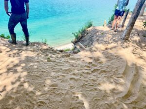 Two people standing on sandy cliff above blue water.