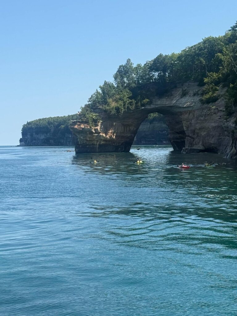 Kayakers near natural rock arch on blue water.