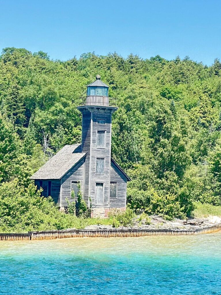 Weathered lighthouse by forested shoreline and blue water.