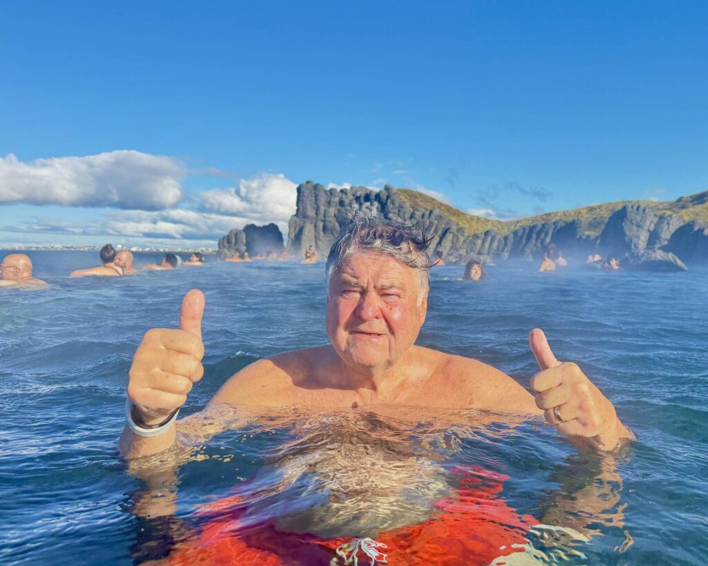 Man enjoying geothermal pool, thumbs up, clear sky.