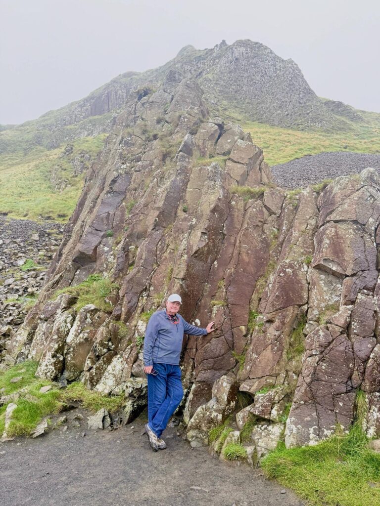Man standing on rocky terrain, grassy hillside background.