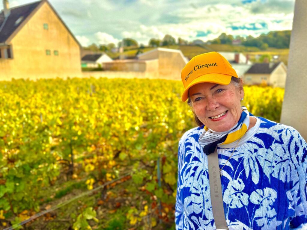 Smiling woman in vineyard, wearing yellow hat.